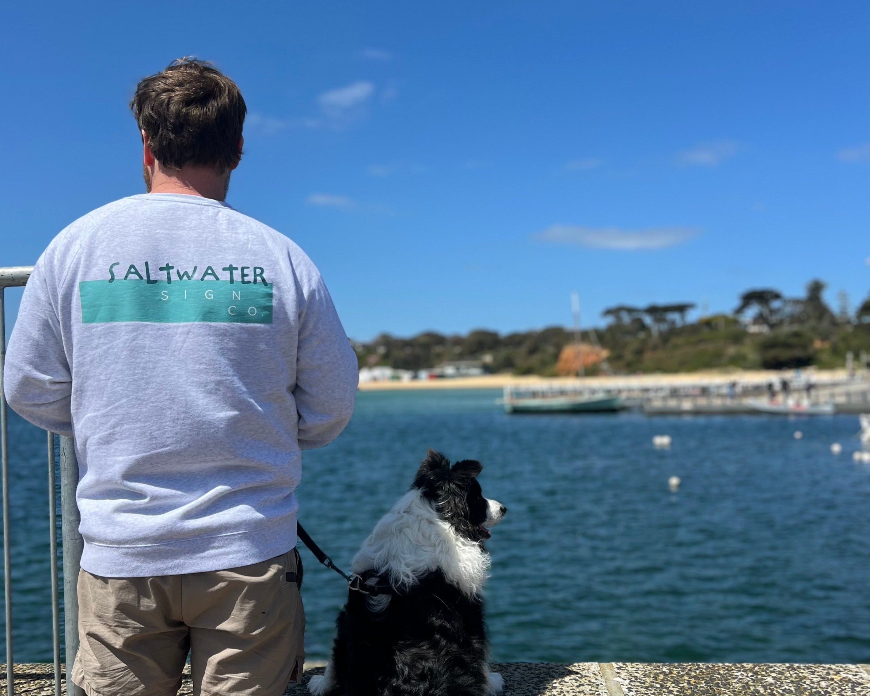 Person wearing a 'Saltwater' shirt with a dog by the Mornington Peninsula waterfront on a clear day